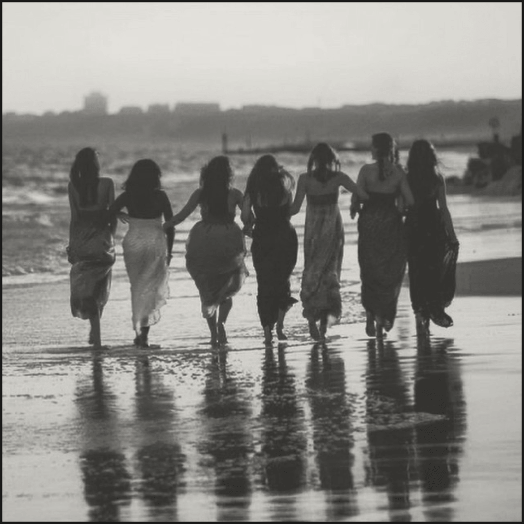 Group of women in dresses walking away on a beach while holding hands.